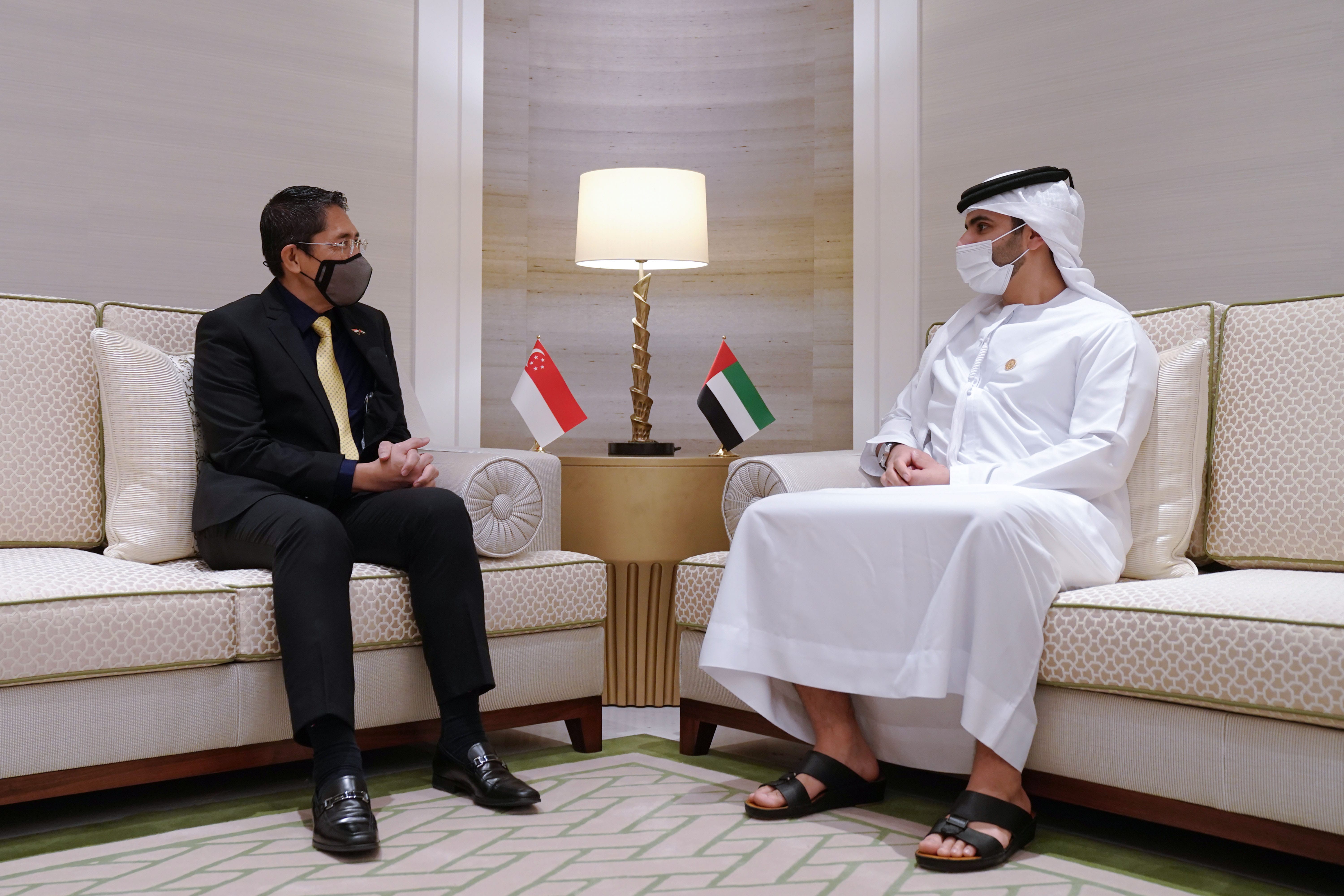 Two men in masks seated on couches, Singapore & UAE flags on table.
